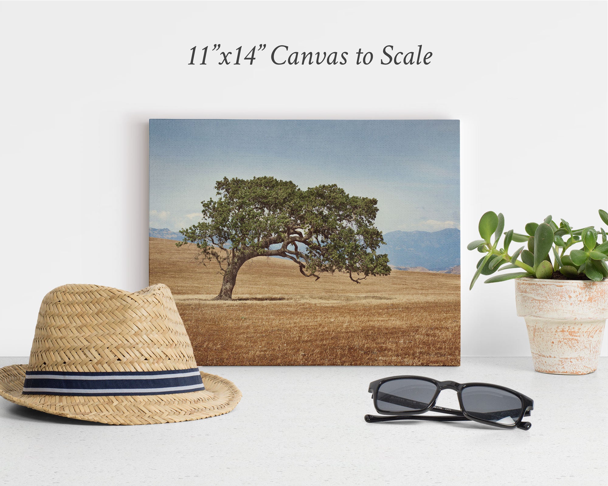 Canvas print of a solitary tree in a field against a mountain backdrop, placed on a surface. A straw hat, sunglasses, and potted plant are arranged in front of the canvas. Text above the canvas reads &quot;11x14 Rustic Canvas Print (Choose from 10+ Designs)&quot; by Offley Green. Perfect for those who appreciate rustic wall art.