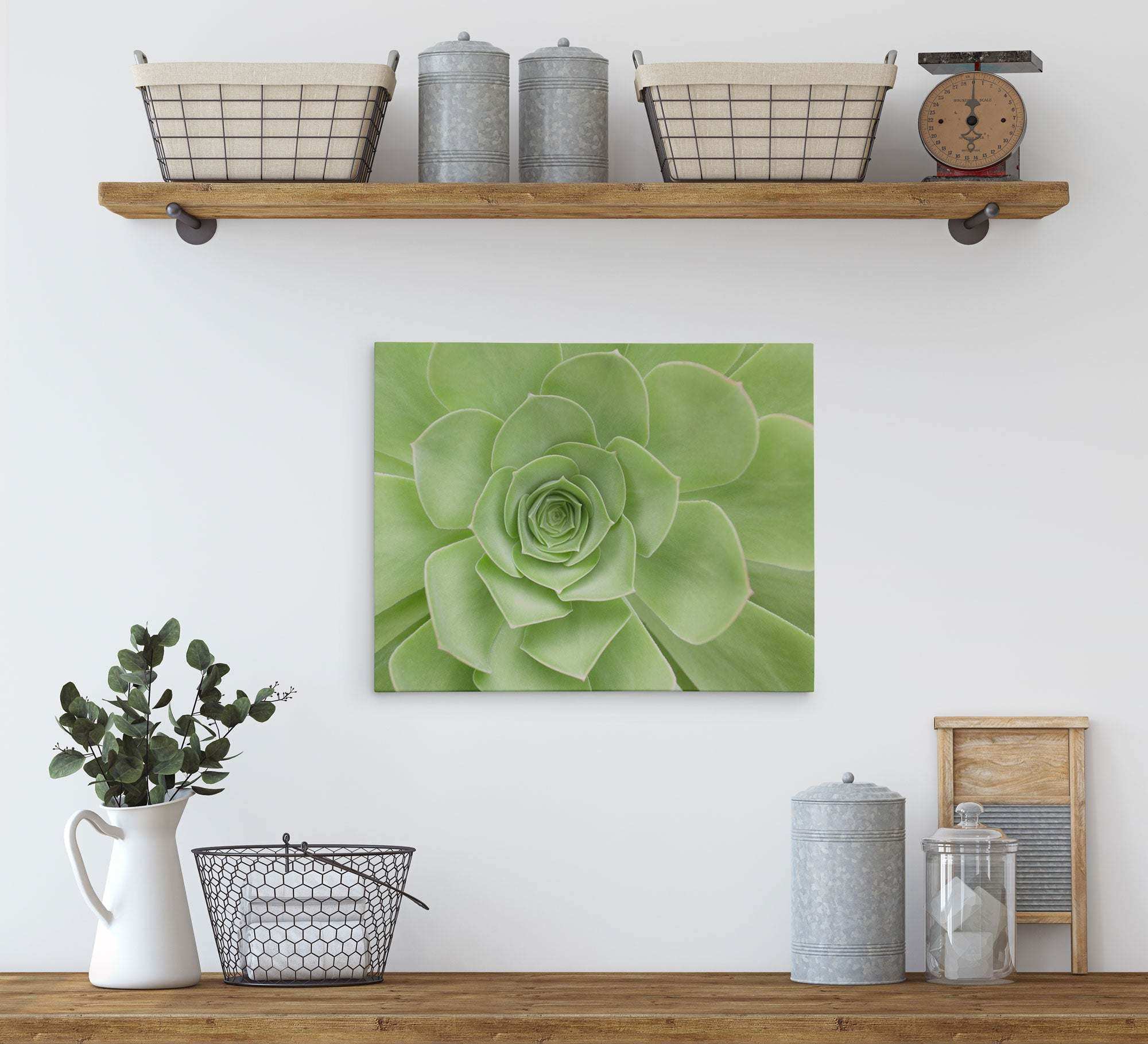 An interior wall shelf holds two wicker baskets and three metal containers, with a vintage scale on the far right. Below, a wooden console displays a white pitcher with green foliage, a wire basket, and additional containers. A green 11x14 Botanical Canvas Print (Choose from 10+ Designs) by Offley Green hangs centered on the wall.