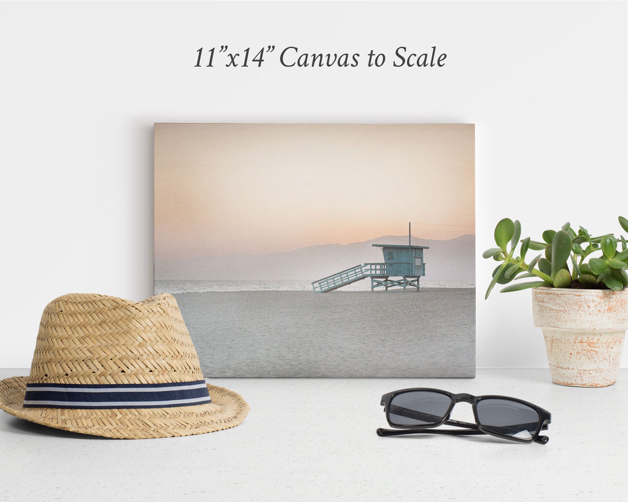 A minimalist beach-themed display features an 11x14 California Canvas Print (Choose from 10+ Designs) by Offley Green of a lifeguard tower at sunset. In front of the canvas are a straw hat with a black band, a pair of black sunglasses, and a small potted plant on a white surface. This captivating wall art evokes serene California prints.