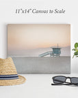 A minimalist beach-themed display features an 11x14 California Canvas Print (Choose from 10+ Designs) by Offley Green of a lifeguard tower at sunset. In front of the canvas are a straw hat with a black band, a pair of black sunglasses, and a small potted plant on a white surface. This captivating wall art evokes serene California prints.