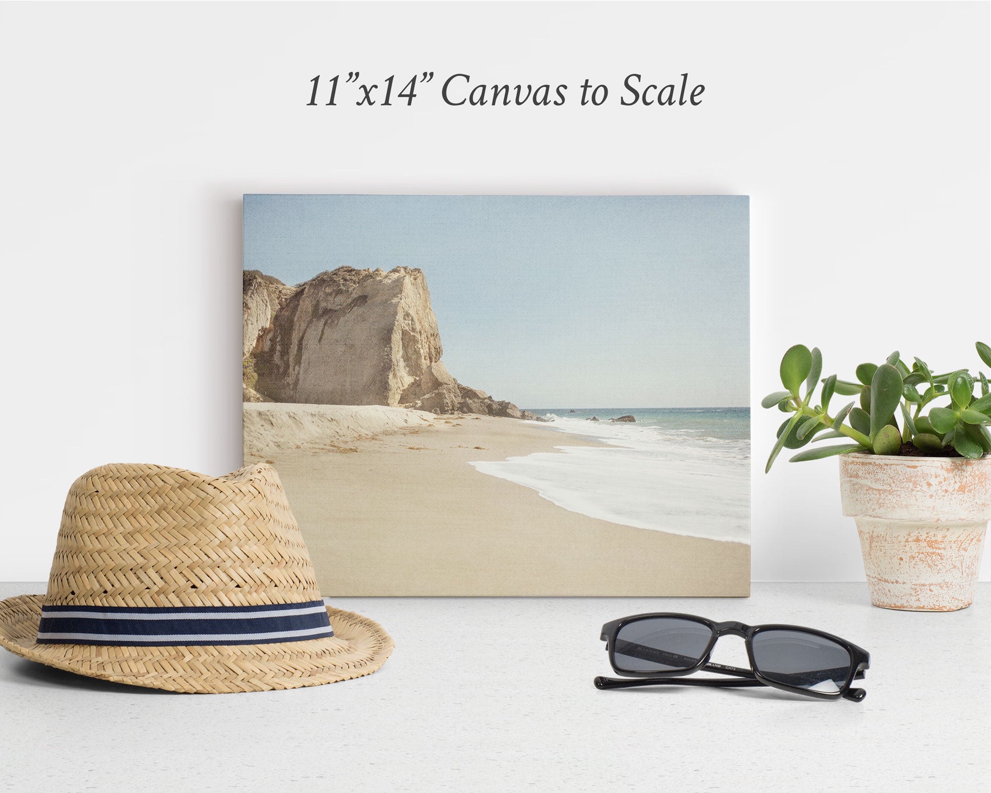 A display setup features an Offley Green 11x14 Coastal Canvas Print, showcasing a beach scene with cliffs and waves. In front of the canvas are a straw hat with a black and white band, a pair of black sunglasses, and a potted plant with green leaves on a white surface, creating the perfect coastal print ambiance.