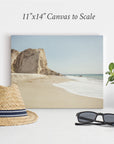 A display setup features an Offley Green 11x14 Coastal Canvas Print, showcasing a beach scene with cliffs and waves. In front of the canvas are a straw hat with a black and white band, a pair of black sunglasses, and a potted plant with green leaves on a white surface, creating the perfect coastal print ambiance.