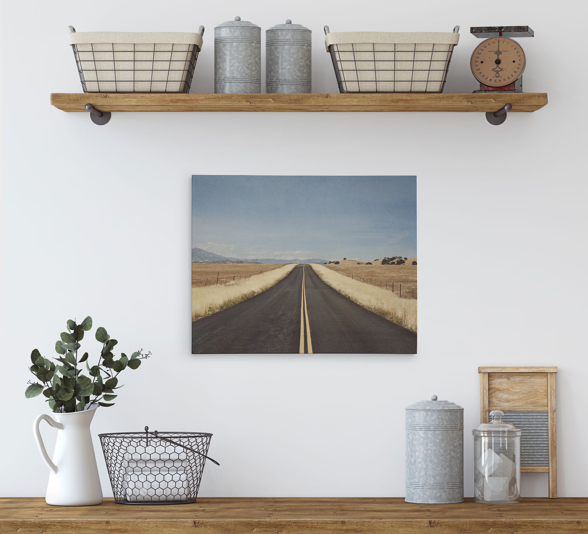 A minimalistic kitchen display with a wooden shelf holding two wicker baskets, metal canisters, and a vintage scale. Below is a countertop with a white pitcher holding greenery, a metal basket, and more canisters. Centered on the wall is an 11x14 Rustic Canvas Print (Choose from 10+ Designs) by Offley Green.