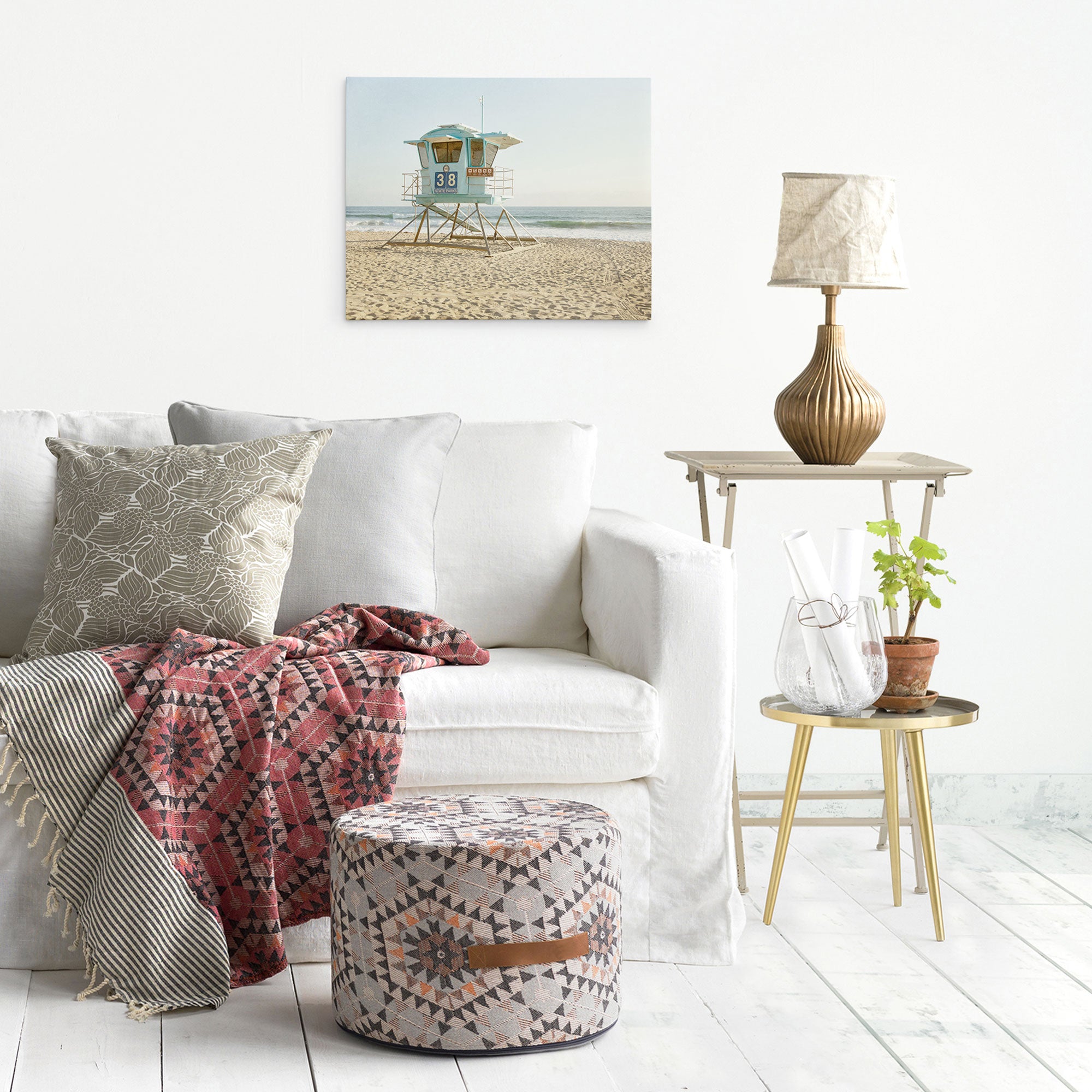 A white living room features a white sofa with patterned and solid cushions. A red and gray patterned blanket is draped over the sofa. Nearby, a round patterned ottoman, side table with a lamp, plant, books, and an Offley Green 16x20 California Canvas Print (Choose from 10+ Designs) complete the cozy setting.