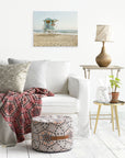 A white living room features a white sofa with patterned and solid cushions. A red and gray patterned blanket is draped over the sofa. Nearby, a round patterned ottoman, side table with a lamp, plant, books, and an Offley Green 16x20 California Canvas Print (Choose from 10+ Designs) complete the cozy setting.