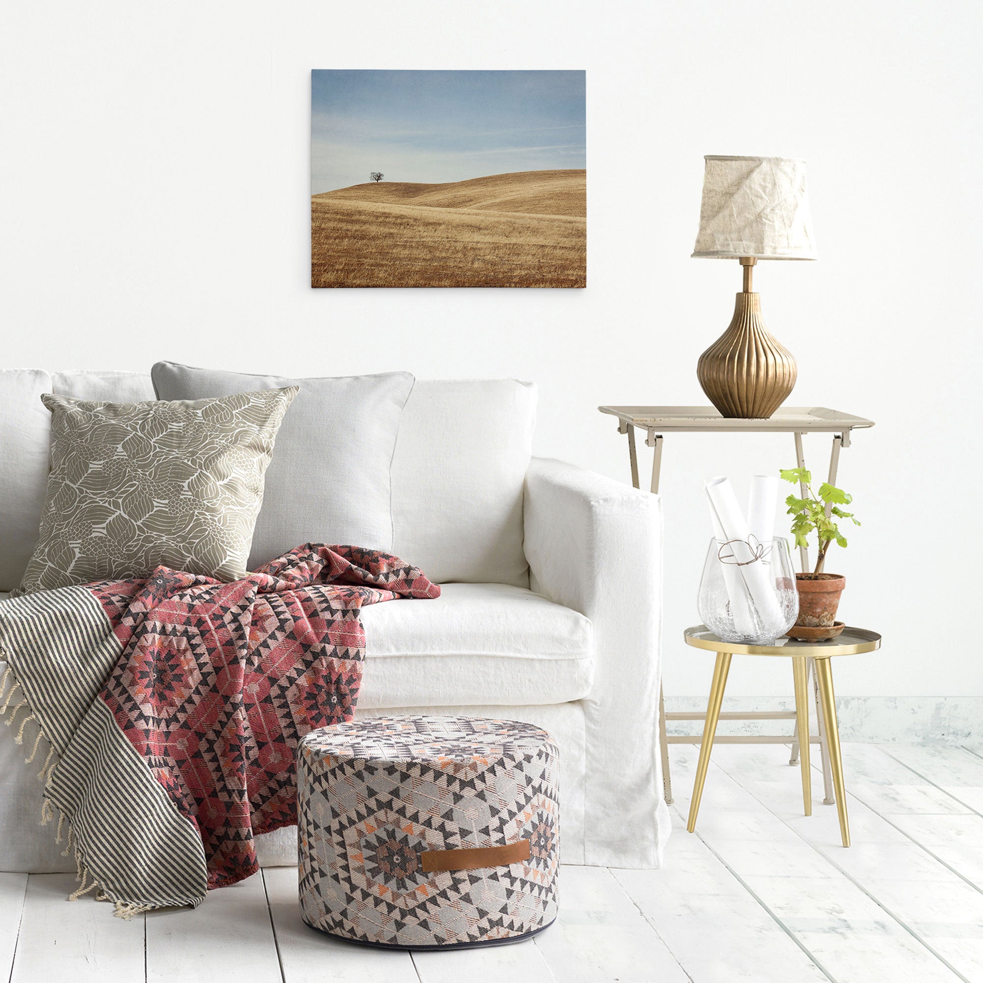 A cozy living room with a white sofa adorned with patterned cushions and a red geometric throw. A small plant, a lamp, and a photo frame sit on a wooden side table. A fabric pouf with geometric patterns lies on the floor. The Offley Green 16x20 Rustic Canvas Print (Choose from 10+ Designs) complements the landscape photo hanging on the white wall.