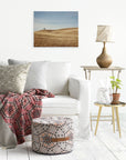 A cozy living room with a white sofa adorned with patterned cushions and a red geometric throw. A small plant, a lamp, and a photo frame sit on a wooden side table. A fabric pouf with geometric patterns lies on the floor. The Offley Green 16x20 Rustic Canvas Print (Choose from 10+ Designs) complements the landscape photo hanging on the white wall.