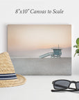 The Offley Green 8x10 California Canvas Print, available in over 10 designs, depicts a beach scene with a lifeguard tower at sunrise or sunset. This gallery wrap wall art rests on a white surface alongside a straw hat adorned with a black and white band, a pair of black sunglasses, and a small potted succulent.