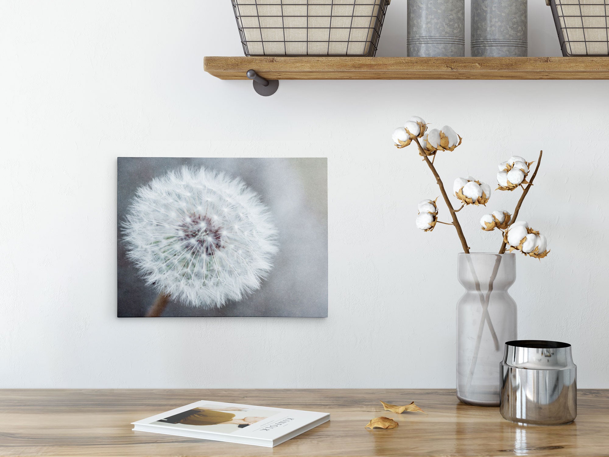 A minimalist interior features a wooden shelf with metal baskets and containers. Below it, a white vase with cotton branches is placed on a wooden surface, accompanied by a metal jar and an open book. An 8x10 Botanical Canvas Print (Choose from 10+ Designs) by Offley Green is mounted on the wall.