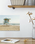 A minimalist interior features a wooden shelf with metal baskets and canisters. Below, a table holds a magazine, a cotton plant in a glass vase, and a metal candle holder. An 8x10 California Canvas Print from Offley Green, depicting one of over ten available designs of a lifeguard tower on a sandy beach, hangs as wall art on the pristine white wall.