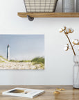 A minimalist interior features a light wooden shelf with metal baskets and two cylindrical containers. Below, an Offley Green 8x10 Coastal Canvas Print featuring a lighthouse on a sandy dune hangs on a white wall. A vase with cotton stems and a candle sit on a wooden table, alongside an open book.