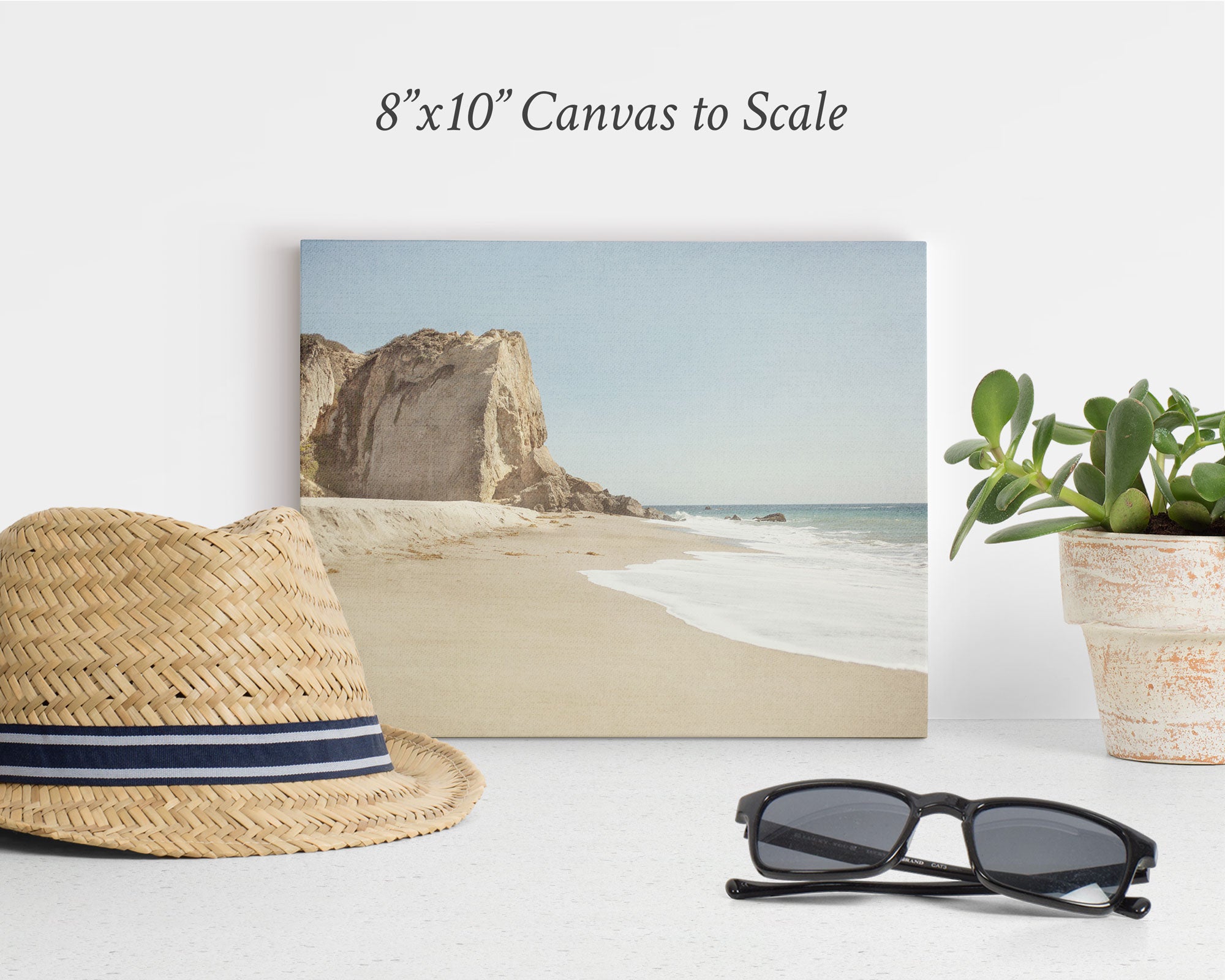 An 8&quot;x10&quot; Coastal Canvas Print from Offley Green, chosen from over 10 available designs, sits on a white surface against a white wall. This beach-themed print beautifully depicts a sandy beach with cliffs in the background. In front, there&#39;s a straw hat with a black and white band, black sunglasses, and a potted plant, making it perfect for your wall art collection.