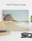 An 8"x10" Coastal Canvas Print from Offley Green, chosen from over 10 available designs, sits on a white surface against a white wall. This beach-themed print beautifully depicts a sandy beach with cliffs in the background. In front, there's a straw hat with a black and white band, black sunglasses, and a potted plant, making it perfect for your wall art collection.