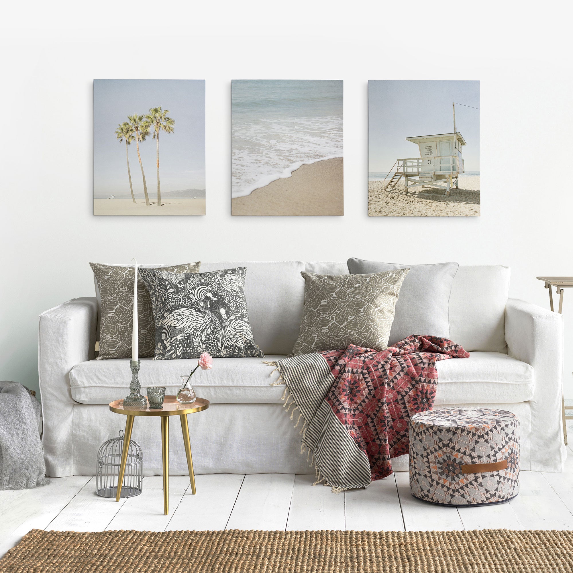 A cozy living room features a white couch with patterned cushions, a textured throw, and a small round table with flowers and candle. Three Offley Green artist-grade beach-themed canvas prints decorate the wall, complemented by a woven pouf and jute rug.