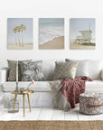 A cozy living room features a white couch with patterned cushions, a textured throw, and a small round table with flowers and candle. Three Offley Green artist-grade beach-themed canvas prints decorate the wall, complemented by a woven pouf and jute rug.