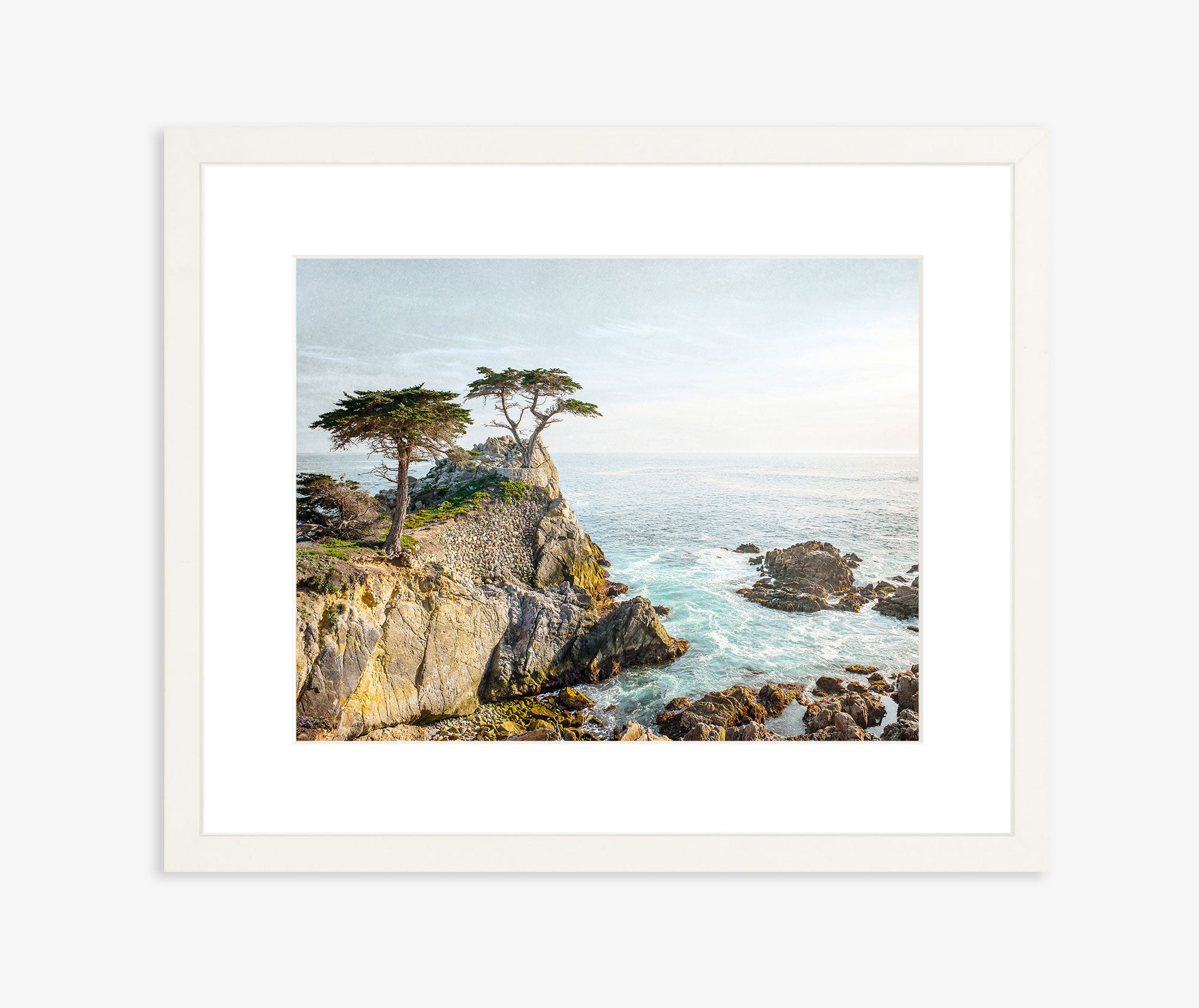 The "Lone Cypress" California Coastal Print by Offley Green showcases a stunning Pebble Beach coastal scene with rocky cliffs and two tall cypress trees poised on the edge. The ocean waves below crash dramatically against the rocks under a clear sky. This beautiful image is printed on archival photographic paper and elegantly framed with a simple, white border.