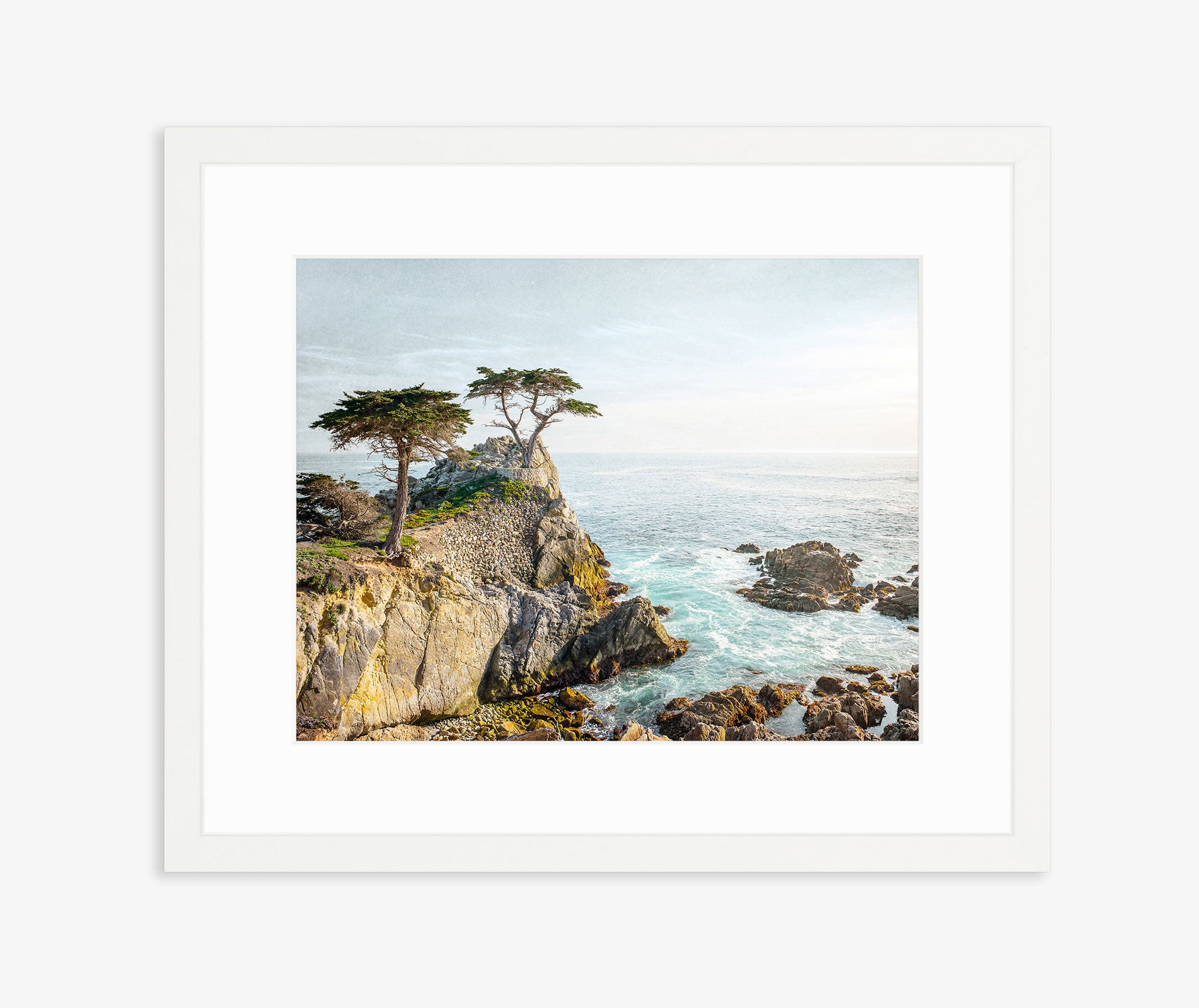 Introducing the California Coastal Print, &#39;Lone Cypress&#39; by Offley Green: a framed photo capturing a serene scene of rocky cliffs topped by two prominent trees, including the iconic solitary cypress tree. This picturesque moment of Pebble Beach features waves crashing against the rocks below, all set against a backdrop of the blue ocean and gentle sky on archival photographic paper.