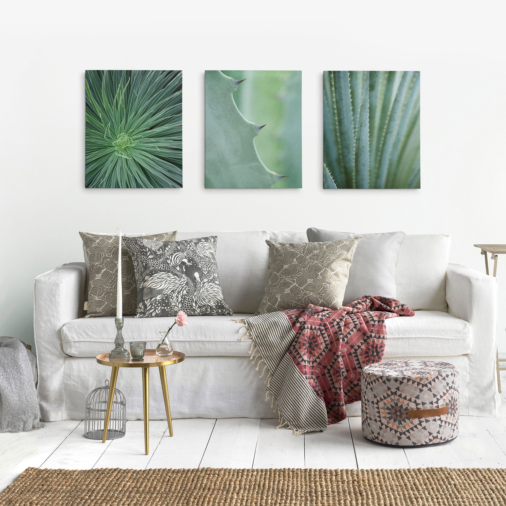 A cozy living room spotlights a white sofa with patterned cushions and a woven throw. Above it are three Offley Green canvas prints featuring botanical art. A round side table, patterned pouf, and textured rug gracefully complete the decor.