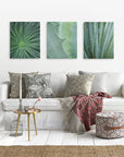 A cozy living room spotlights a white sofa with patterned cushions and a woven throw. Above it are three Offley Green canvas prints featuring botanical art. A round side table, patterned pouf, and textured rug gracefully complete the decor.