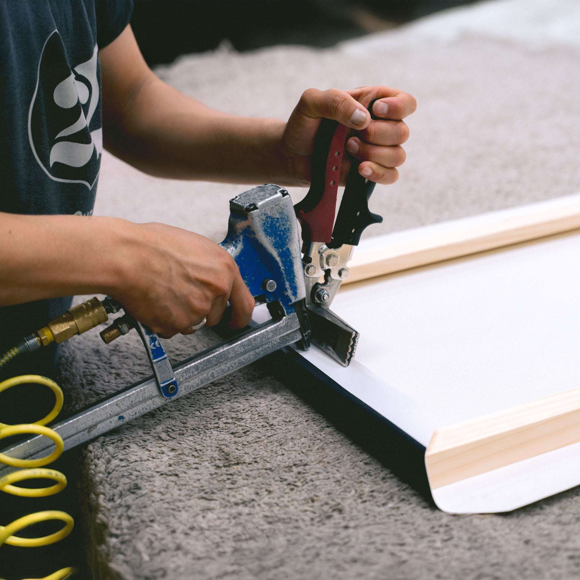 Close-up of a person using a staple gun to affix an Offley Green 24x30 Rustic Canvas Print (Choose from 10+ Designs) to a wooden frame. The person&#39;s hands grip the stapler, and the canvas is partially wrapped around the frame. Various tools and materials suggest a workshop or art studio setting, ideal for creating rustic wall art.