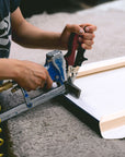 An individual using a pneumatic staple gun to secure fabric onto a wooden frame, designed for Offley Green's Set of 4 Framed Canvas (choose from multiple combinations) in botanical or coastal styles. The wooden frame sits on a soft, textured surface as the person's hands grip the staple gun and ensure the fabric is pulled tight for perfectly framed canvas prints.