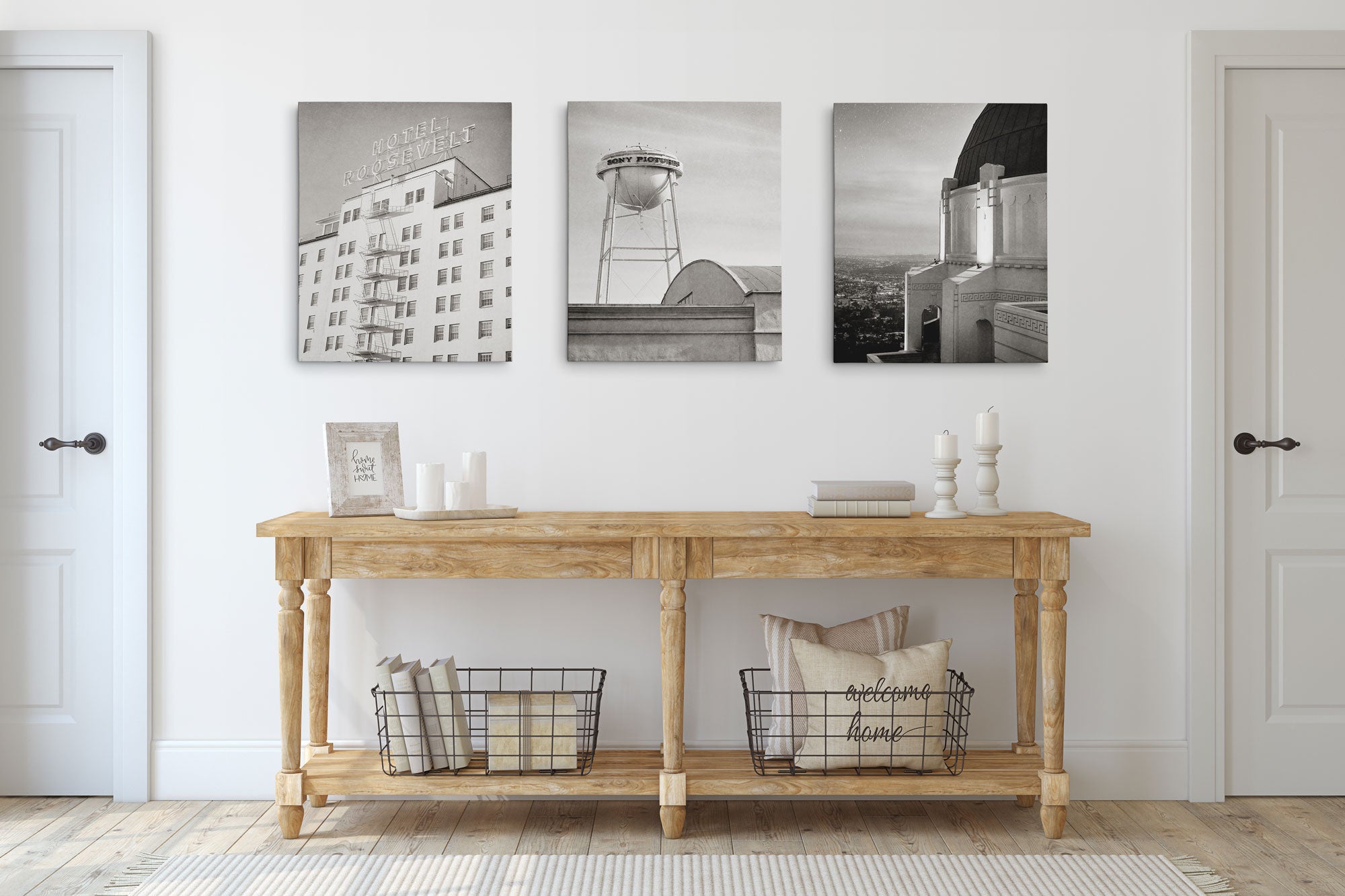 The room features a wooden console table with candles, books, and baskets holding pillows. Above it, the Offley Green Set of 3 Canvas displays black and white photos of a hotel, water tower, and observatory. White walls and doors elegantly frame the space.