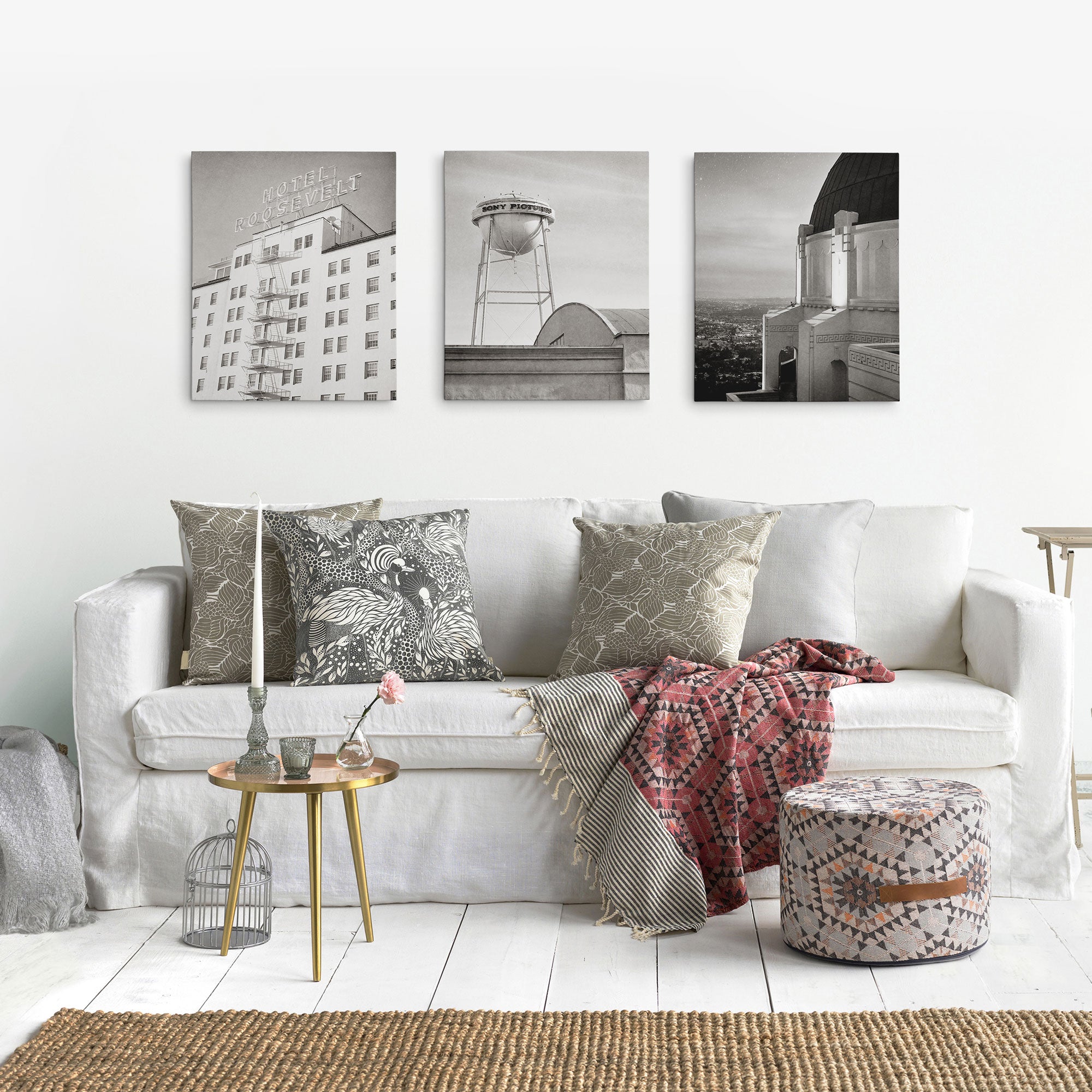 A cozy living room showcases a white sofa with patterned pillows, a red throw, and a woven stool. A round side table holds flowers and candles, while natural light highlights three cityscape prints from the Offley Green Set of 3 Canvas hanging above.