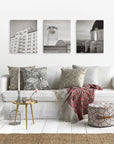 A cozy living room showcases a white sofa with patterned pillows, a red throw, and a woven stool. A round side table holds flowers and candles, while natural light highlights three cityscape prints from the Offley Green Set of 3 Canvas hanging above.