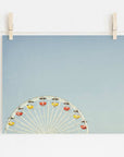 A photograph pinned by wooden clothespins on a string, depicting the top half of a ferris wheel with colorful cabins at Santa Monica Pier, set against a plain light blue background - Blue Minimalist Wall Decor, 'Ferris Blue' by Offley Green.