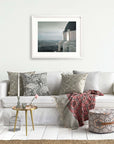 A minimalist living room featuring a white sofa adorned with patterned pillows, a small metallic side table with decor, a woven pouf, and an Offley Green archival photograph print of Griffith Observatory ('The Sky At Night') hanging on the wall.