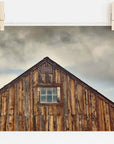 A photograph of a rustic farmhouse with a pitched roof and a single window, pinned up on white archival photographic paper by wooden clothespins. Dark clouds loom in the background. Offley Green's 'Old Barn at Bodie' Farmhouse Rustic Print is a stunning addition to any decor.