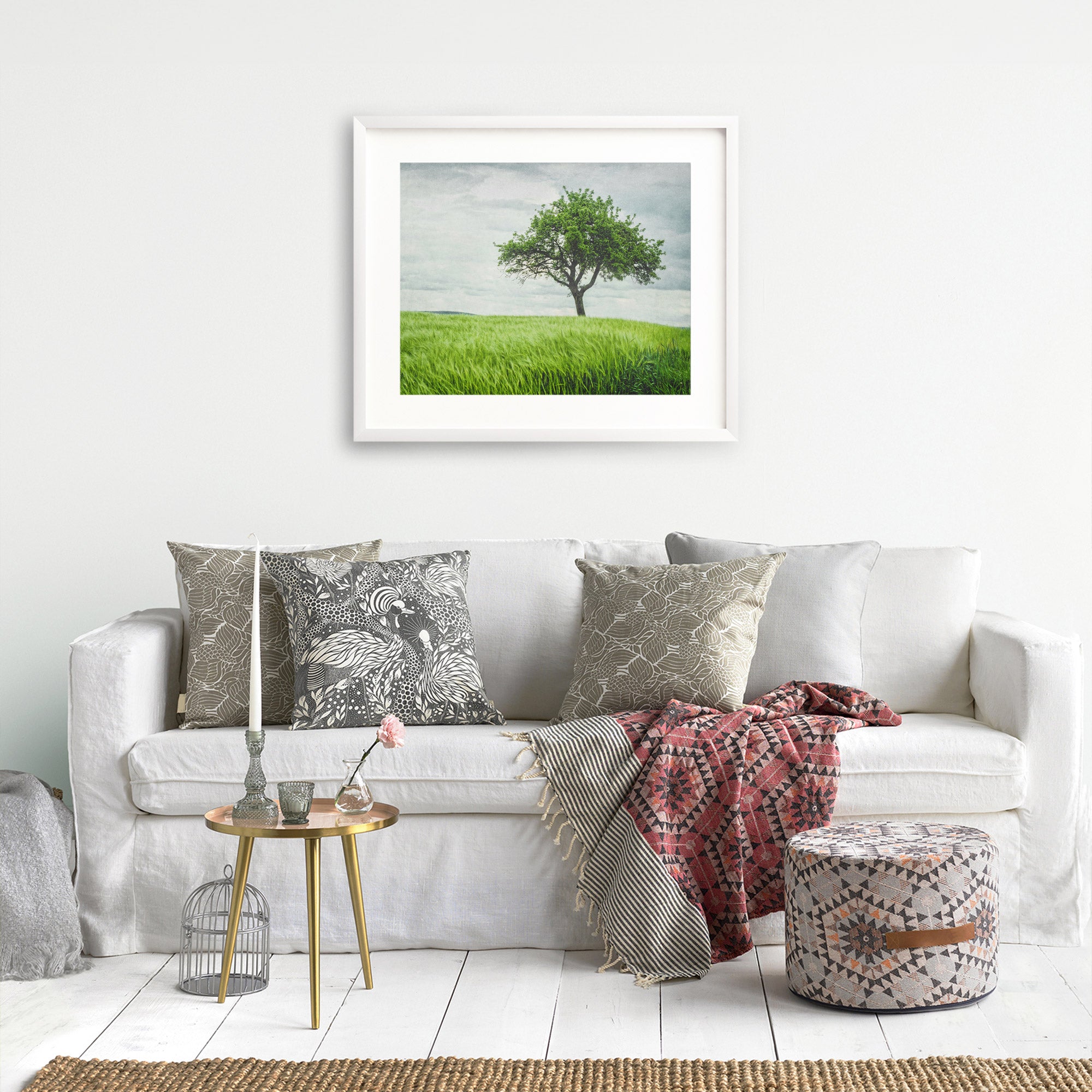 A cozy living room features a white sofa adorned with patterned cushions and a red patterned throw. A small round table with a vase and candle holder sits in front. A framed Offley Green Rustic Countryside Print, 'Tree in a Field' hangs on the wall, and a pouf rests on a woven rug.