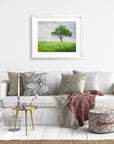 A cozy living room features a white sofa adorned with patterned cushions and a red patterned throw. A small round table with a vase and candle holder sits in front. A framed Offley Green Rustic Countryside Print, 'Tree in a Field' hangs on the wall, and a pouf rests on a woven rug.