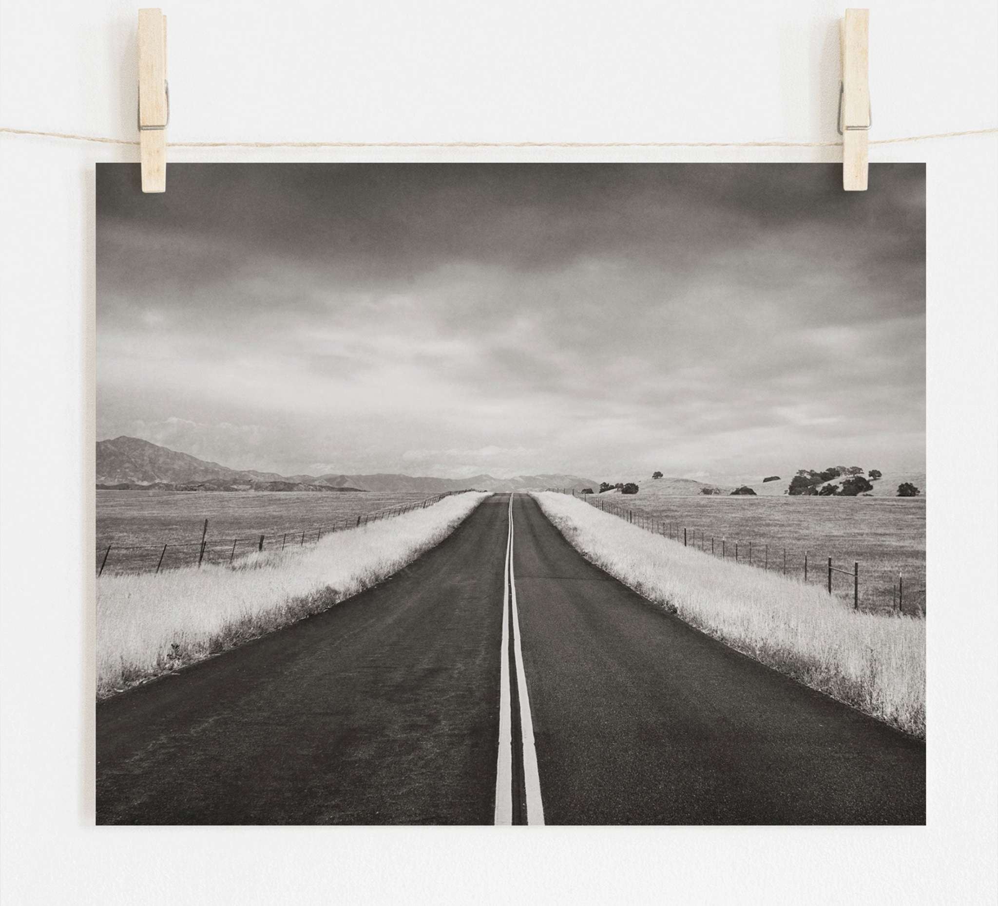 Offley Green&#39;s Black and White Rural Landscape Art, &#39;American Road Trip&#39;, printed on archival photographic paper, features an empty road extending into the distance under a cloudy sky, flanked by fields and mountains in sepia tones.