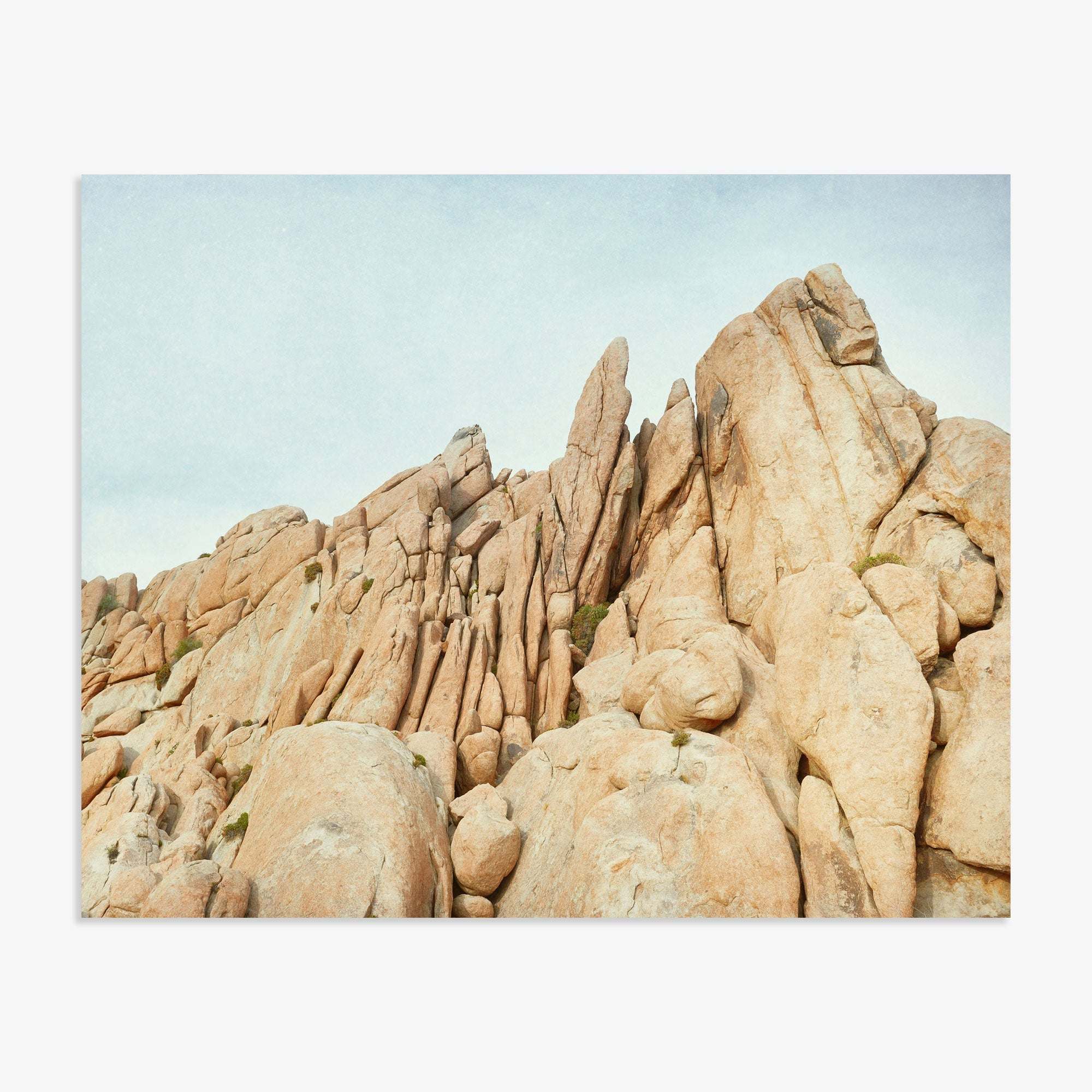 Rugged and textured rock formations in Joshua Tree rise steeply under a clear sky, with sharp peaks and large, rounded boulders covering the landscape. This scene is captured beautifully in the Offley Green &#39;Joshua Rocks&#39; print.