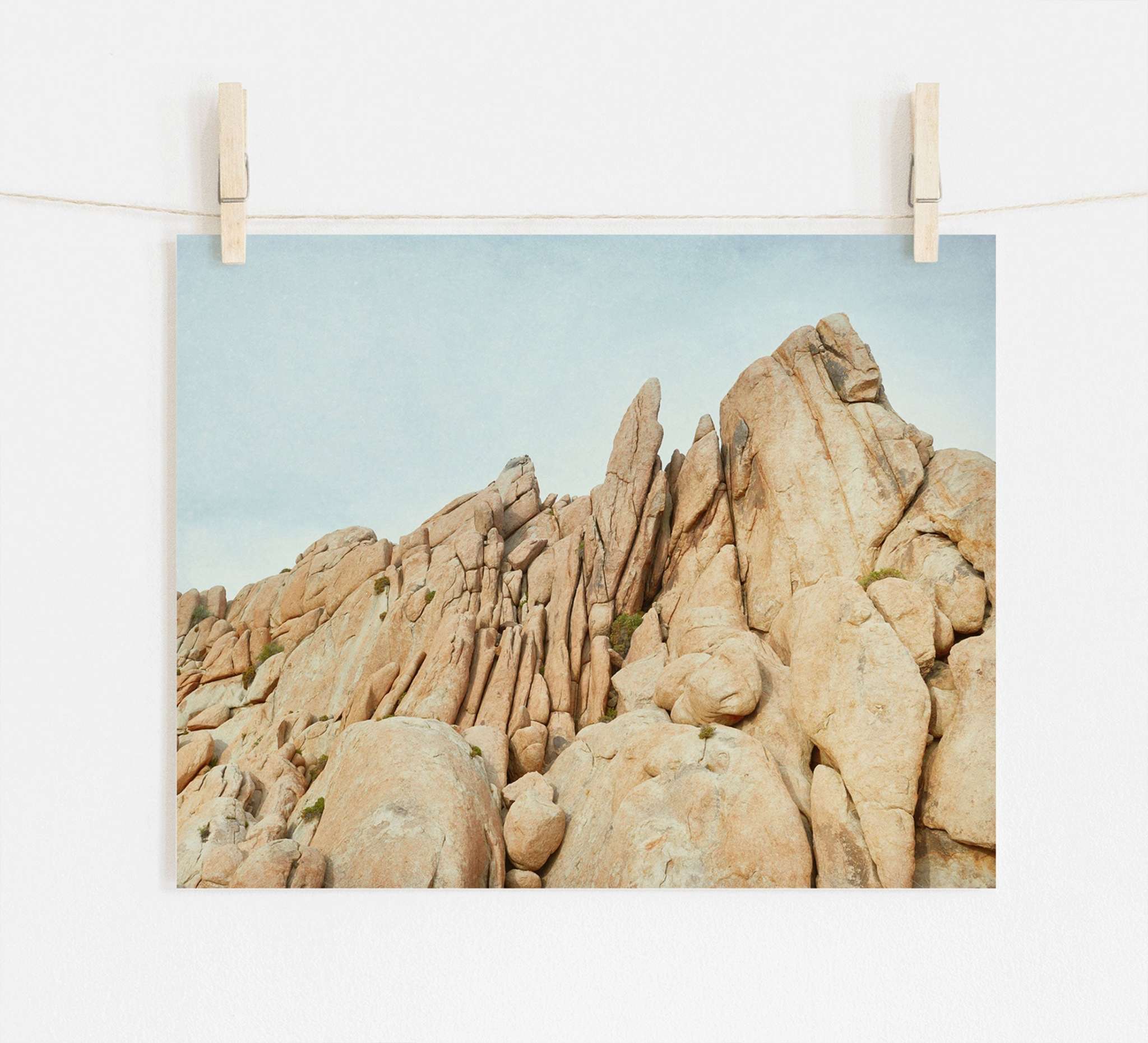 A photo of rugged, rocky cliffs with pointed formations, displayed hanging from two wooden clothespins on a string against a white wall, printed on archival photographic paper. 

Replace with: Offley Green&#39;s Joshua Tree Print, &#39;Joshua Rocks&#39;