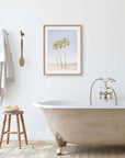 A minimalist bathroom featuring a clawfoot bathtub with golden fixtures, a wooden stool with bath items, and an unframed Offley Green California Venice Beach Print, 'Three Palms' on the wall next to hanging towels.