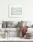 A cozy living room setup with a white Offley Green sofa adorned with the Coastal Beach Print in California 'Surf and Sun' patterned cushions, a small gold side table, a wicker ottoman with a pink flower on top, and a gray cage on the floor.