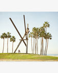 A large, abstract metal sculpture comprising angular beams, located on a grassy area with tall palm trees near Venice Beach. A person on a bicycle is visible in the foreground. This is the Offley Green Venice Beach Landmark Sculpture, 'V is for Venice'.