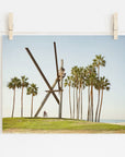 A photograph of the Venice Beach Landmark Sculpture, 'V is for Venice', by Offley Green, surrounded by tall palm trees, printed on archival photographic paper, hangs on a white wall displayed with wooden clips on a string. In the background, a person on a.