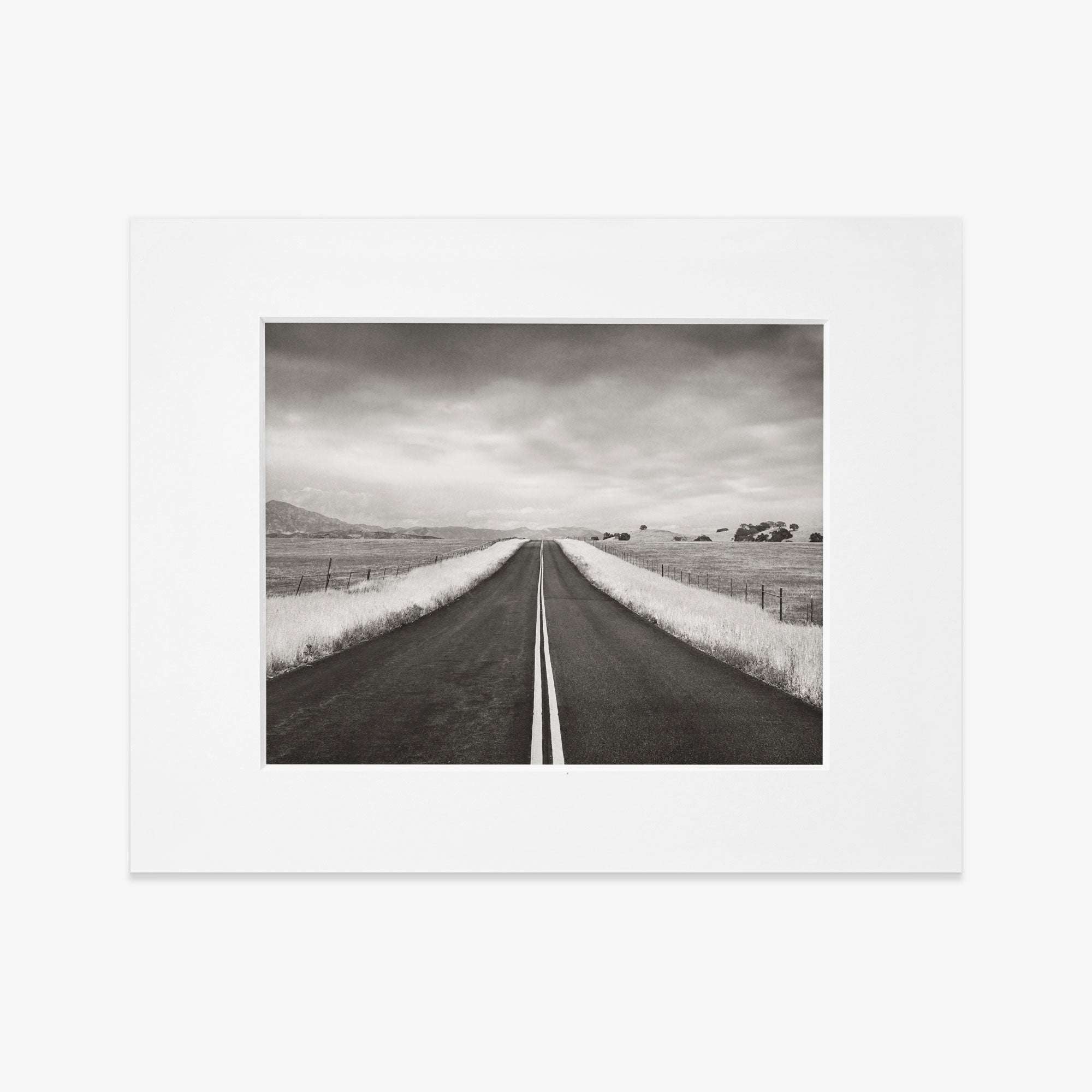 Black and white photograph of a straight road framed by a white border, printed on archival photographic paper, leading through a flat rural landscape under a cloudy sky. This is the Black and White Rural Landscape Art, &#39;American Road Trip&#39; from Offley Green.