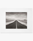 Black and white photograph of a straight road framed by a white border, printed on archival photographic paper, leading through a flat rural landscape under a cloudy sky. This is the Black and White Rural Landscape Art, 'American Road Trip' from Offley Green.