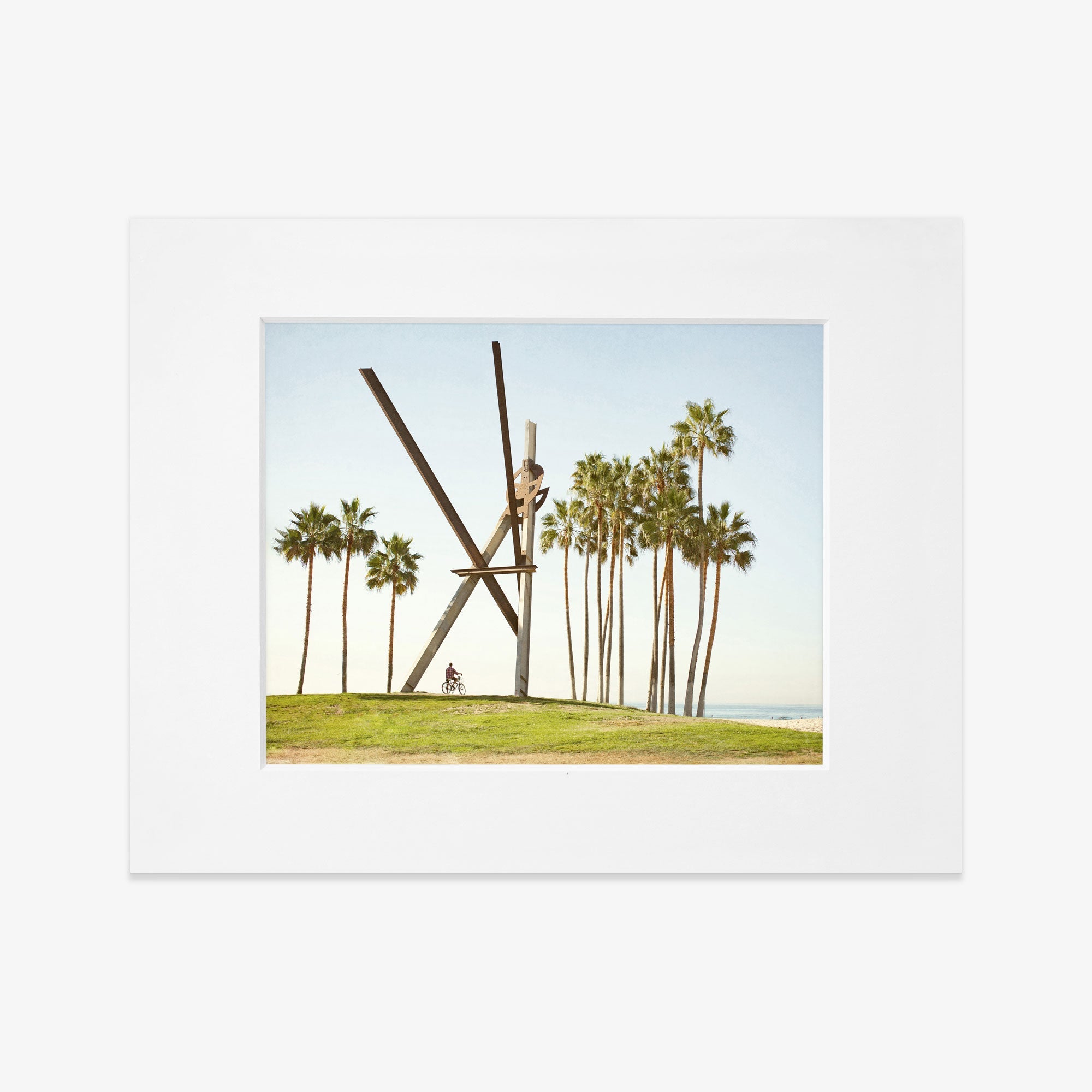 A framed photograph of the Venice Beach Landmark Sculpture, &#39;V is for Venice&#39; by Offley Green, surrounded by tall palm trees under a clear blue sky. A person on a bicycle is visible near the sculpture, captured on archival photographic paper.