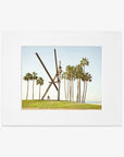 A framed photograph of the Venice Beach Landmark Sculpture, 'V is for Venice' by Offley Green, surrounded by tall palm trees under a clear blue sky. A person on a bicycle is visible near the sculpture, captured on archival photographic paper.
