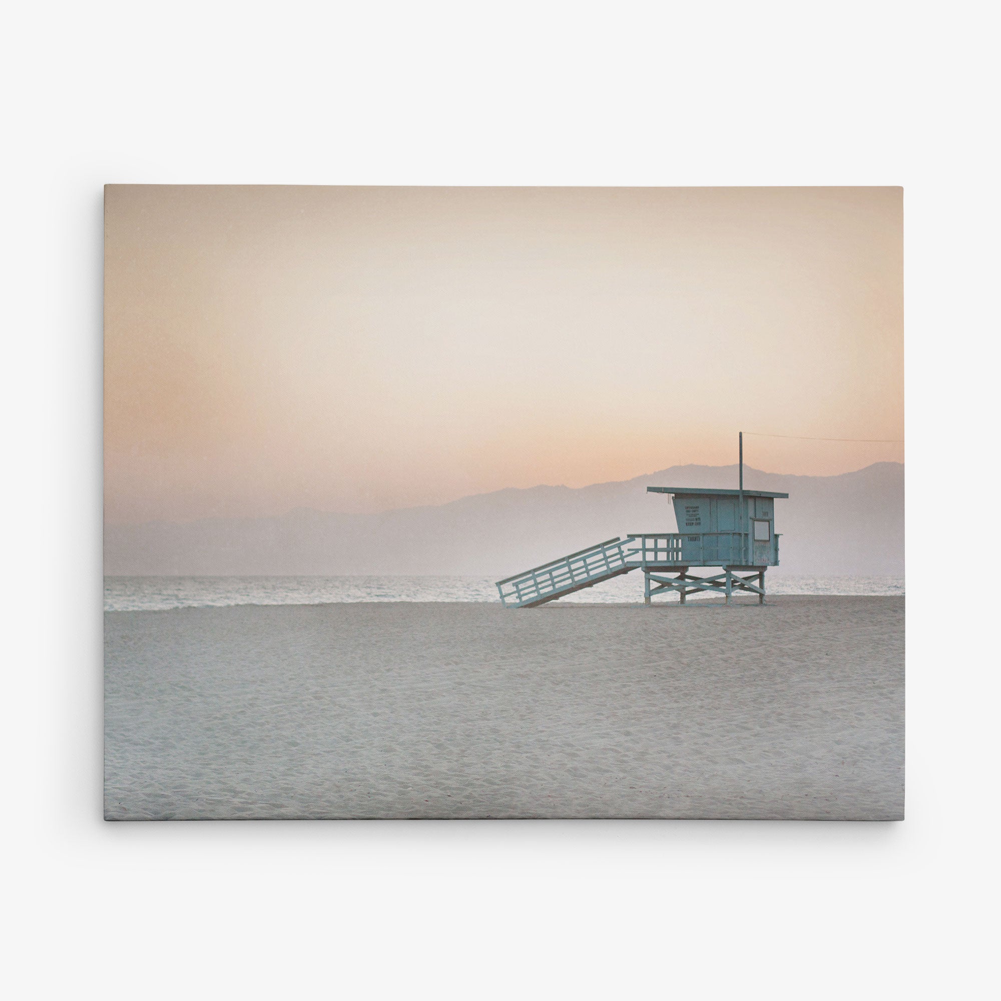 A solitary lifeguard tower stands on a sandy beach at sunset, with soft orange and pink hues in the sky and distant mountains fading into the horizon. This tranquil scene, perfect for Offley Green&#39;s 11x14 California Canvas Print (Choose from 10+ Designs), captures the serene beauty of the coastal landscape.