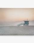 A solitary lifeguard tower stands on a sandy beach at sunset, with soft orange and pink hues in the sky and distant mountains fading into the horizon. This tranquil scene, perfect for Offley Green's 11x14 California Canvas Print (Choose from 10+ Designs), captures the serene beauty of the coastal landscape.