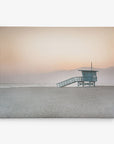 A solitary lifeguard tower stands on a deserted, sandy beach under a pastel sunset sky. The hues of pink, orange, and purple fade into the horizon with distant, silhouetted mountains. The calm ocean lies beyond the lifeguard tower, reflecting the serene ambiance—perfect for Offley Green's 24x30 California Canvas Print (Choose from 10+ Designs) as canvas gallery wrap wall art.