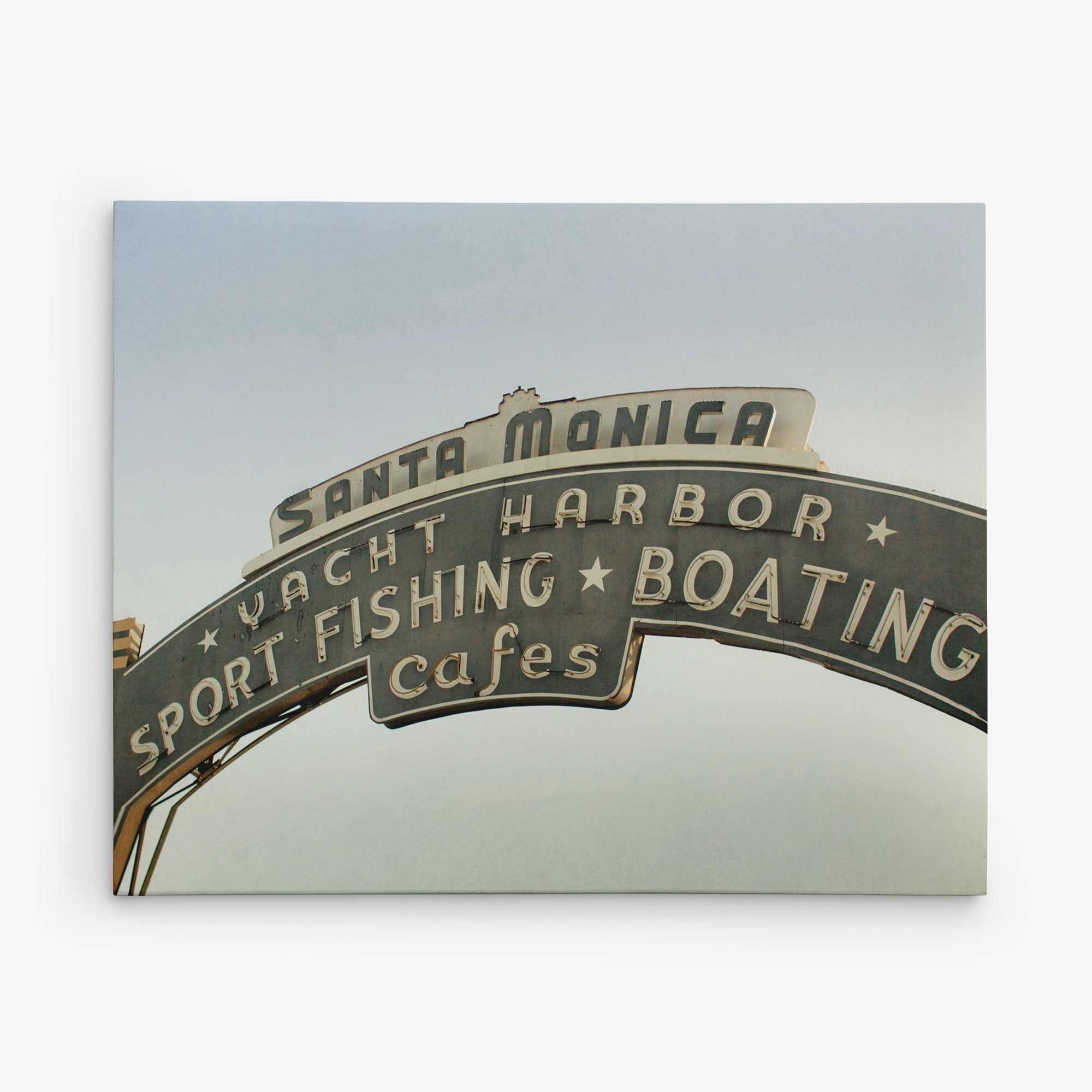 A 30x40 California Canvas Print from Offley Green featuring a curved sign displaying &quot;Santa Monica&quot; at the top, with &quot;Yacht Harbor&quot; below it and &quot;Sport Fishing Boating Cafes&quot; in smaller text. The lighted sign is set against a clear sky background, making it an ideal choice to elevate your wall art collection. Choose from over 10 designs to find your perfect match.