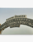 A 30x40 California Canvas Print from Offley Green featuring a curved sign displaying "Santa Monica" at the top, with "Yacht Harbor" below it and "Sport Fishing Boating Cafes" in smaller text. The lighted sign is set against a clear sky background, making it an ideal choice to elevate your wall art collection. Choose from over 10 designs to find your perfect match.