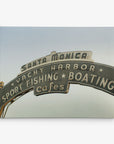 An arch-shaped sign reads "Santa Monica Yacht Harbor Sport Fishing Boating Cafes." The sign is photographed from a low angle against a clear sky, perfect for Offley Green's 11x14 California Canvas Print (Choose from 10+ Designs).